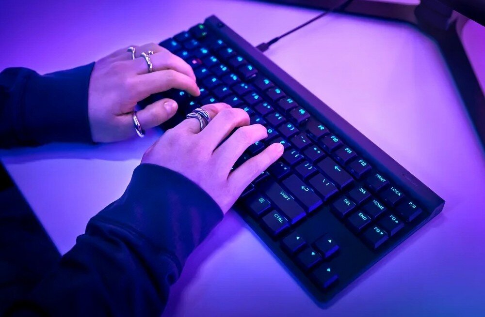 Osoba pisze na czarnej klawiaturze z podświetleniem RGB. Klawiatura jest podłączona kablem, a scena jest oświetlona fioletowym światłem.