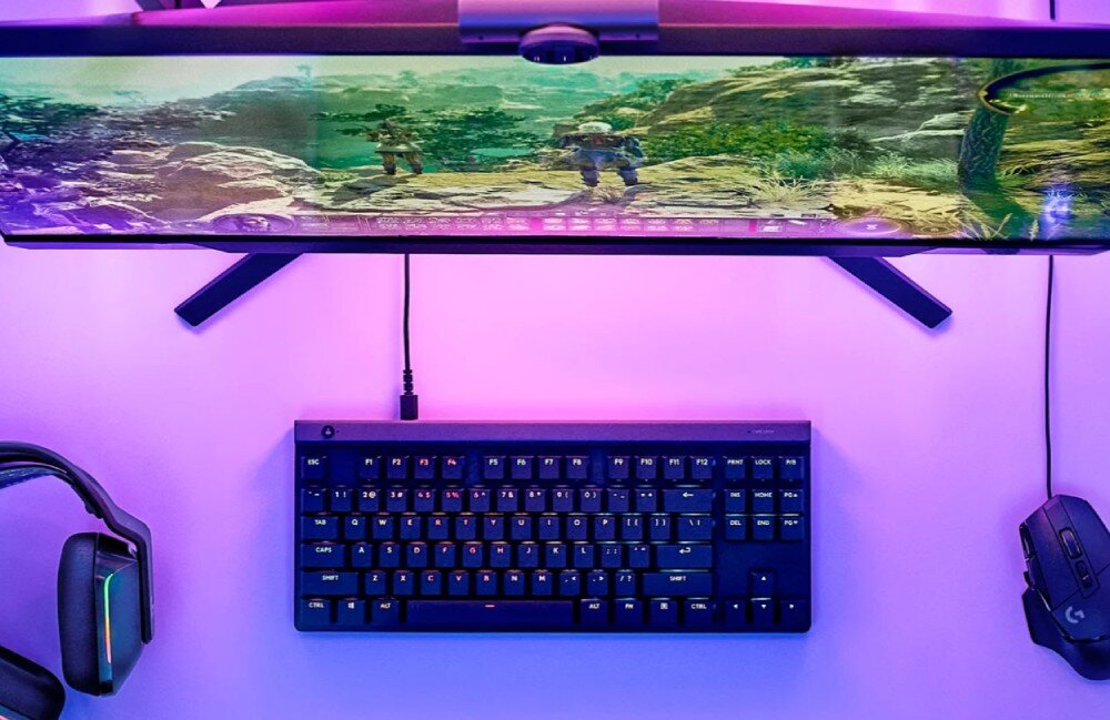 Na biurku znajduje się czarna klawiatura mechaniczna z podświetleniem RGB. Obok widoczne są słuchawki i mysz komputerowa, a na monitorze wyświetlana jest gra komputerowa.