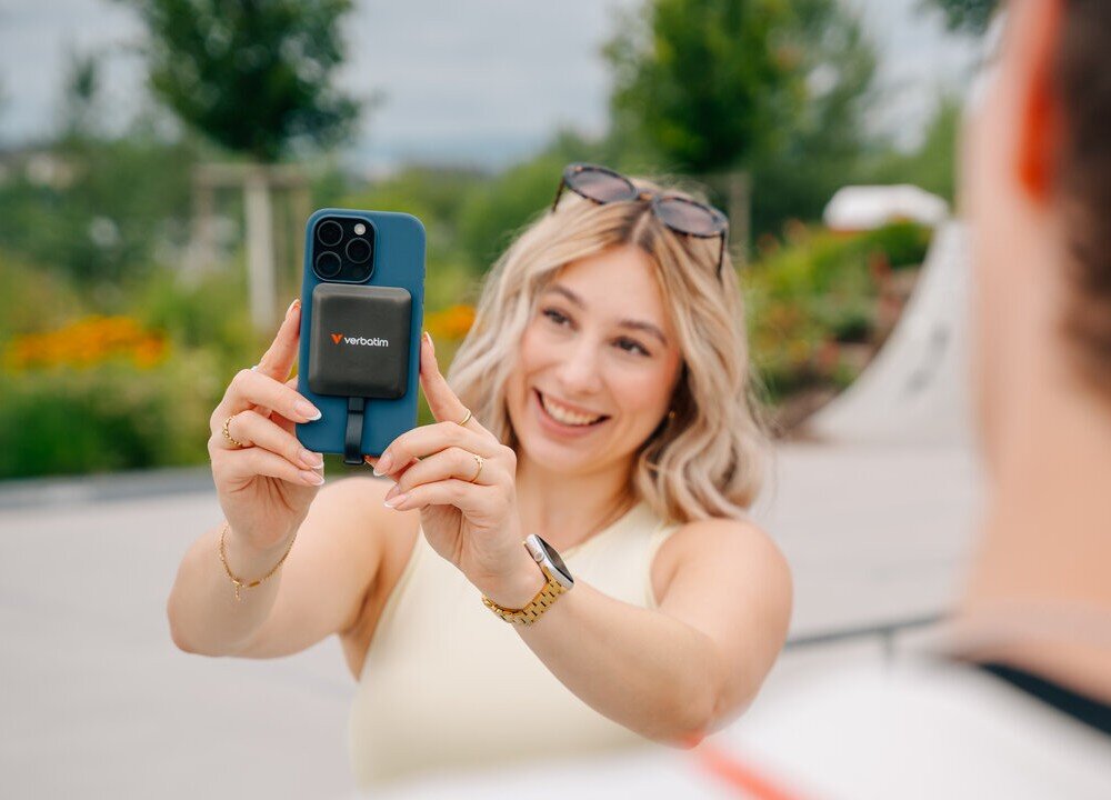 Dysk VERBATIM SnapBack Uśmiechnięta kobieta trzyma oburącz smartfon z przymocowanym dyskiem SSD Verbatim, skierowany w stronę niewidocznego rozmówcy, opis