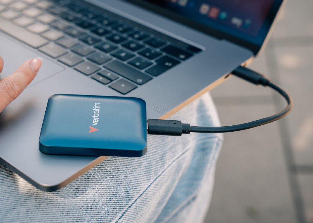 Dysk VERBATIM SnapBack Zbliżenie na dysk SSD Verbatim leżący na kolanach w jeansach, połączony krótkim kablem z portem USB-C otwartego laptopa, pojemność, przeznaczenie