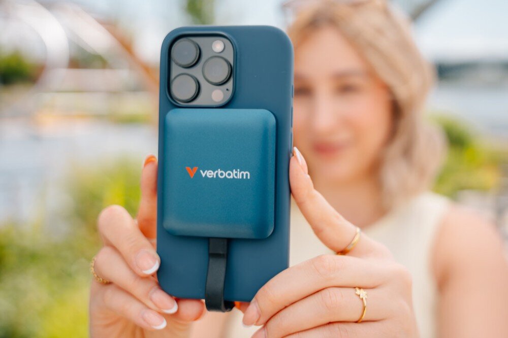Dysk VERBATIM SnapBack Uśmiechnięta kobieta trzyma oburącz smartfon z przymocowanym dyskiem SSD Verbatim, skierowany w stronę niewidocznego rozmówcy, opis