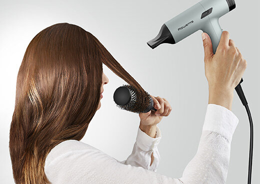 Kobieta suszy długie włosy suszarką ROWENTA HY7120, jednocześnie modeluje je okrągłą szczotką.