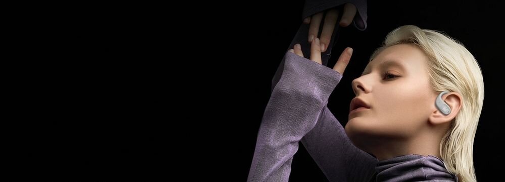 Osoba z jasnymi włosami nosi kremowe słuchawki douszne. Tło jest czarne, a osoba ma na sobie fioletowy strój z długimi rękawami.