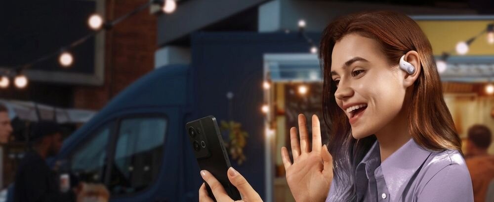 Osoba trzymająca smartfon i nosząca kremowe słuchawki powietrzne. W tle widoczne są rozmyte światła i pojazd.