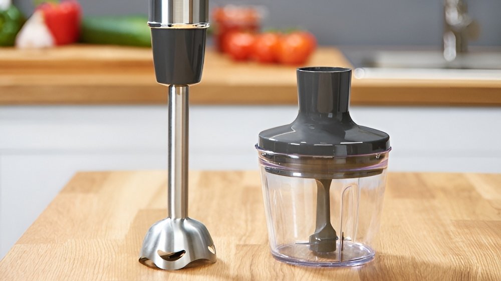 Na drewnianym blacie kuchennym znajduje się metalowy blender ręczny oraz przezroczysty pojemnik z szarym wieczkiem i ostrzem wewnątrz. W tle widoczne są rozmyte elementy kuchni, takie jak zlew i warzywa.