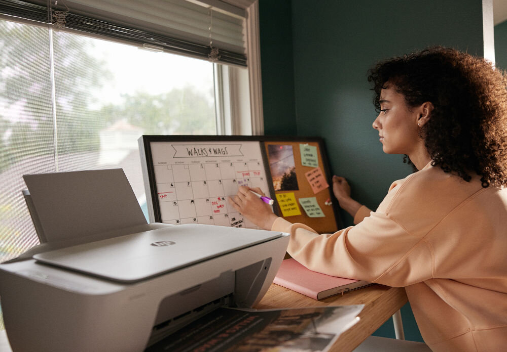Urządzenie wielofunkcyjne HP DeskJet 2821e Druk w kolorze, Wi-Fi, Instant Ink prezentacja urządzenia na biurku obok tablica do markerów i kobieta pisząca po niej za biurkiem okno kompatybilność Windows macOS różne środowiska