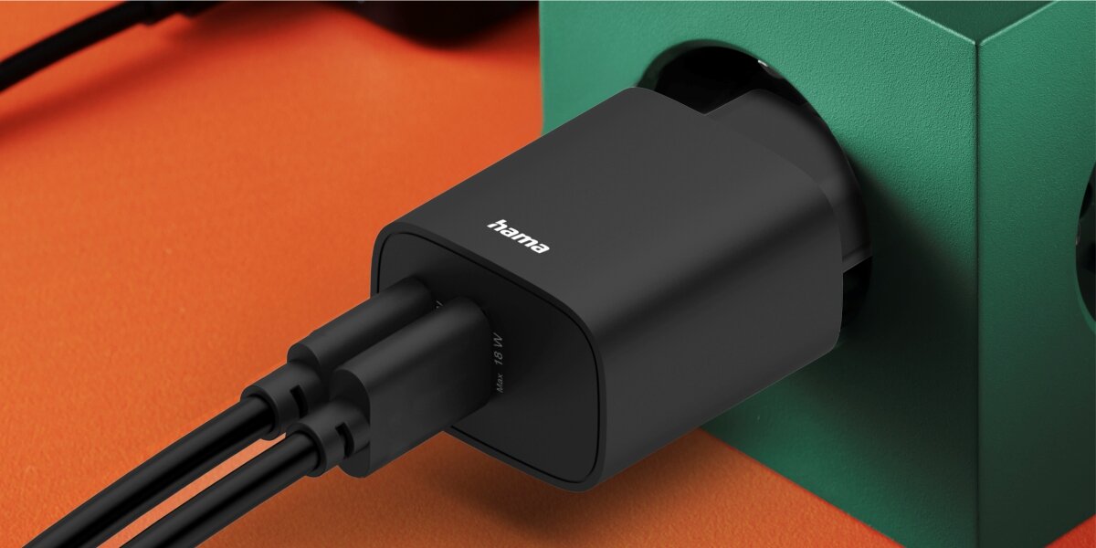 Ładowarka Hama z dwoma podłączonymi kablami USB wpięta do gniazdka elektrycznego w zielonej obudowie.