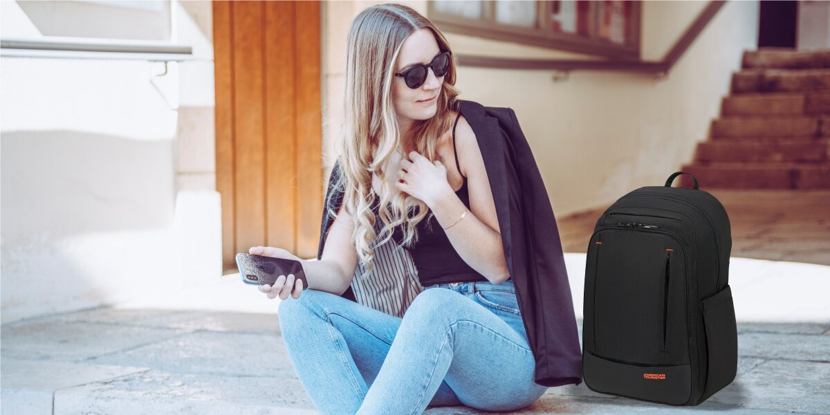 Plecak na laptopa AMERICAN TOURISTER Urban Groove 155520-1041 15.6 cala Kobieta siedząca na schodach z telefonem, obok której stoi czarny plecak na laptopa American Tourister.