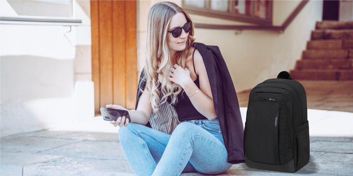Plecak na laptopa AMERICAN TOURISTER Urban Groove 155519-1041 15.6 cala Czarny Kobieta w okularach przeciwsłonecznych siedząca na schodach z telefonem w ręku i czarnym plecakiem American Tourister obok niej