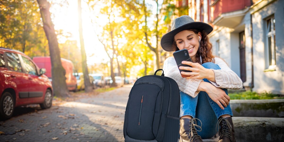 Plecak na laptopa AMERICAN TOURISTER Urban Groove 155517-1265 17.3 cala Granatowy Uśmiechnięta kobieta w kapeluszu siedząca na murku z telefonem i Granatowy plecak na laptopa American Tourister na pierwszym planie