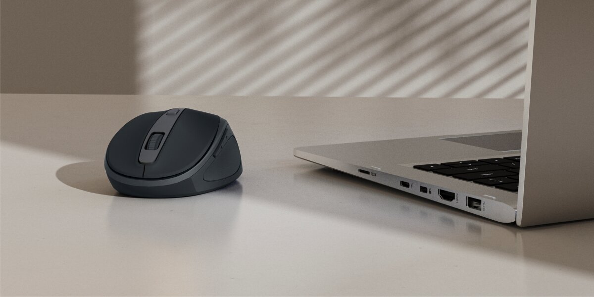 Bezprzewodowa mysz komputerowa Hama w kolorze grafitowym/antracytowym, leżąca na biurku obok otwartego laptopa (notebooka) ze srebrną obudową. Scena oświetlona bocznym światłem, sugerująca wygodną pracę biurową.