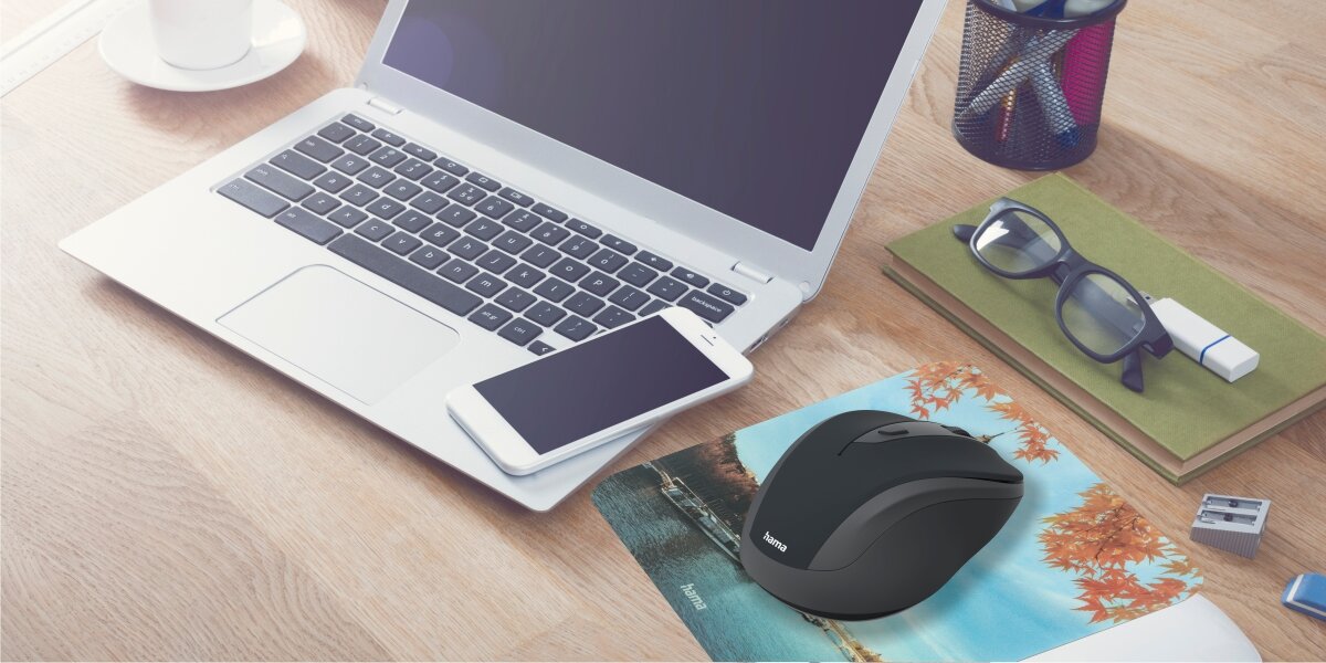 Bezprzewodowa mysz komputerowa Hama w kolorze czarno-szarym, leżąca na podkładce z motywem jesiennym, obok otwartego laptopa i smartfona. Scena na drewnianym biurku z dodatkami biurowymi, sugerująca ergonomiczne stanowisko pracy.