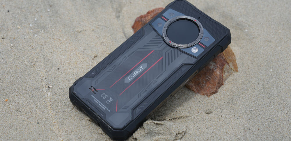 Smartfon CUBOT King Kong 11 5G 16/256GB 6.72 120Hz Czarny Pancerny smartfon Cubot leżący tyłem na piaszczystej plaży obok kamienia, ukazujący wytrzymałą obudowę i okrągły wyświetlacz pomocniczy