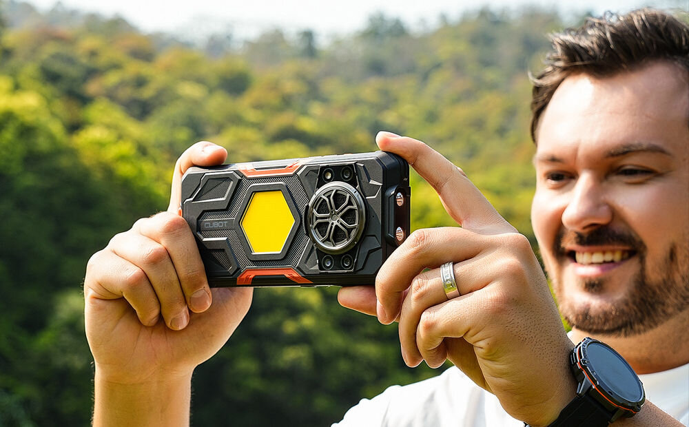 Smartfon CUBOT KING KONG Power 5 16/128GB 6.88 120Hz Czarny Uśmiechnięty mężczyzna trzyma pancerny smartfon Cubot w pozycji poziomej, fotografując lub nagrywając wideo. Widoczna jest tylna obudowa z kamerami, dużą żółtą wkładką i kratką głośnika. Tło to zalesiony krajobraz