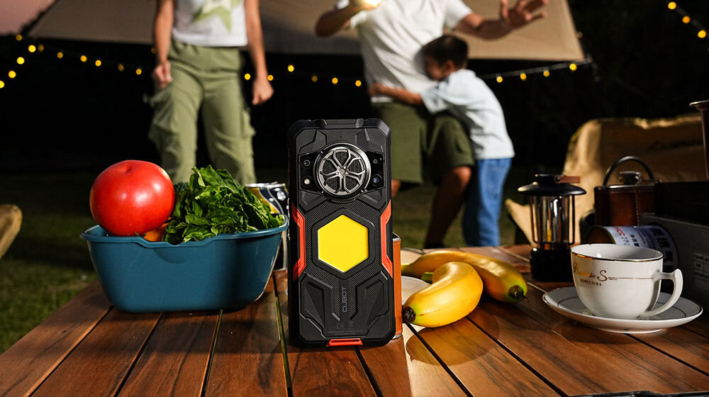Smartfon CUBOT KING KONG Power 5 16/128GB 6.88 120Hz Czarny Pancerny smartfon Cubot leży na drewnianym stole na kempingu. W tle, niewyraźnie, widać ludzi przy namiocie i dekoracyjne lampki. Obok smartfona leżą owoce i naczynia