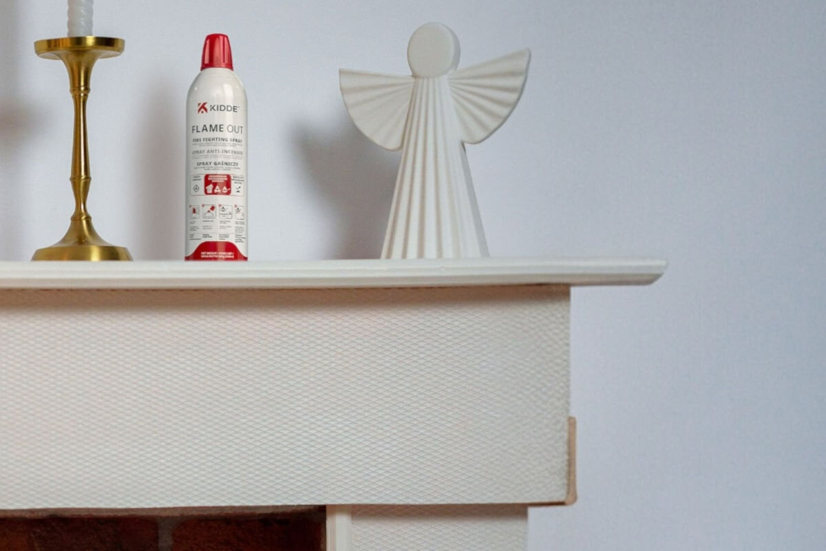 SPRAY GAŚNICZY KIDDE FLAMEOUT (500 ML), szafka, schowek, lekki, niewielki, Spray Kidde FlameOut stojący na beżowej półce obok figurki anioła i złotego świecznika.