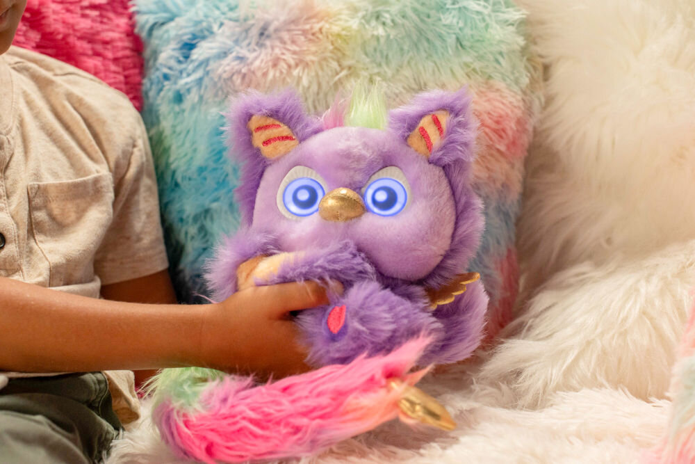 Zabawka interaktywna FURLINGS Furlittles Curiously Cute Gryffin 26117 Dziecko delikatnie trzyma interaktywną maskotkę Gryffin z kolorowym ogonem, siedząc wśród miękkich, kolorowych poduszek.