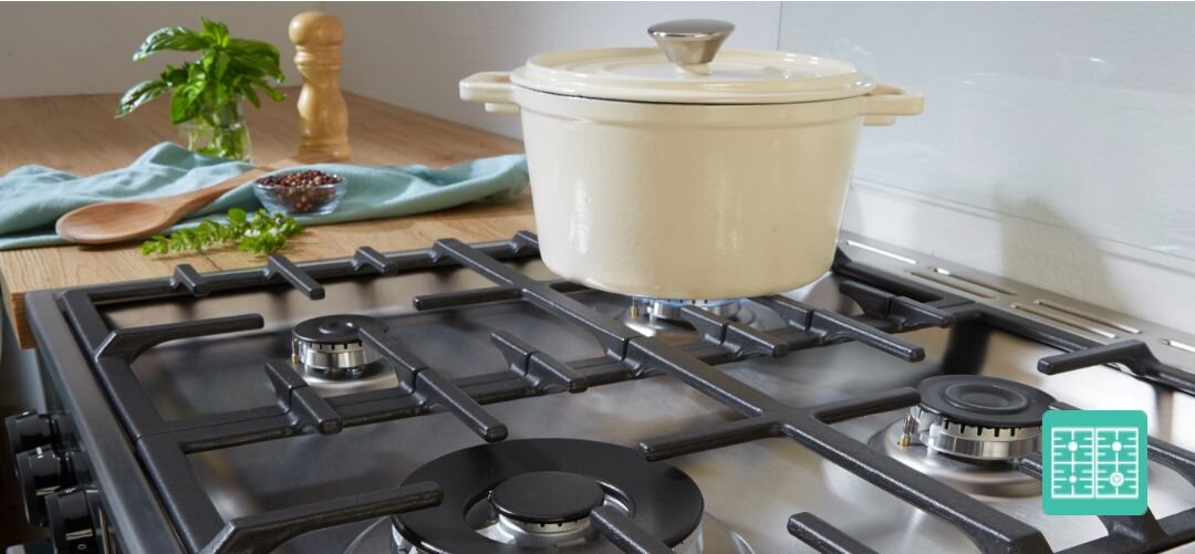 KUCHNIA GORENJE GKS6C71WA Widok na płytę gazową z pięcioma palnikami oraz masywnymi, żeliwnymi rusztami, na środkowym palniku stoi kremowy garnek z pokrywką, a w tle na drewnianym blacie leżą drewniana łyżka, przyprawy i doniczka z ziołami ruszty ProGrids stabilne odporne