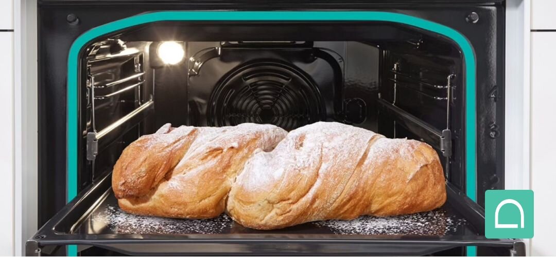 KUCHNIA GORENJE GK5C42WH Dwie duże, rumiane chałki posypane cukrem pudrem pieką się na blasze w oświetlonym wnętrzu piekarnika, którego obrys zaznaczono turkusową linią podkreślającą specjalny kształt komory