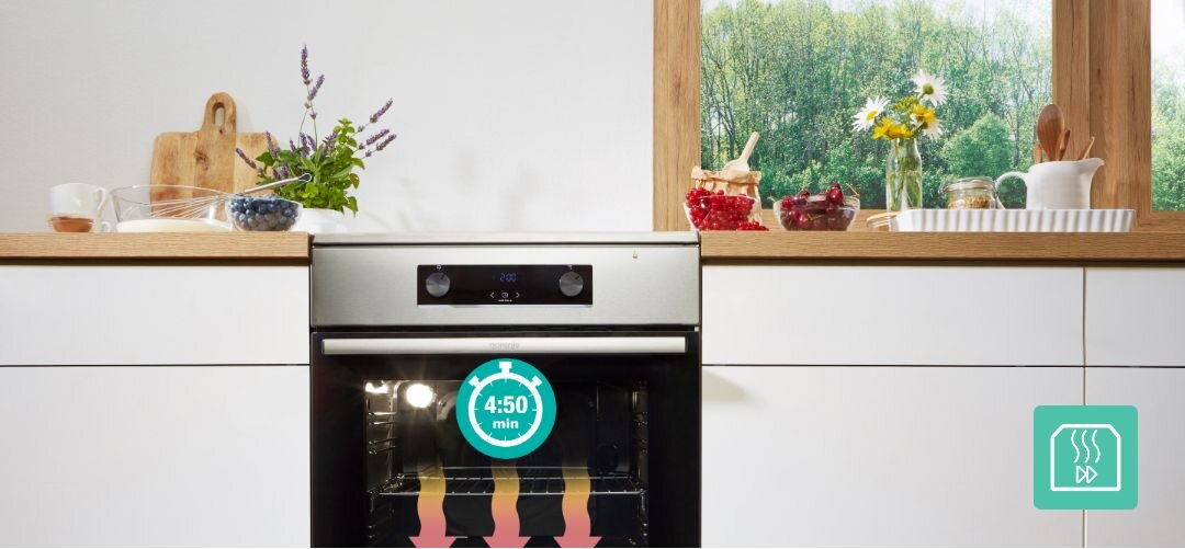 KUCHNIA GORENJE GK5C42WH Zabudowany piekarnik Gorenje w jasnej kuchni stoi pod dużym oknem z widokiem na zieleń, na blacie wokół ustawiono naczynia, owoce i zioła, a na drzwiach piekarnika widoczna jest grafika zegara z czasem 4:50 min oraz ikoną pary, podkreślająca funkcję szybkiego nagrzewania z wykorzystaniem gorącego powietrza szybkie nagrzewanie krótki czas