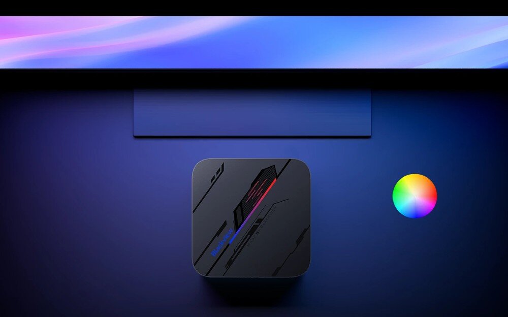 Komputer BLACKVIEW MP100 Pro Widok z góry na małe czarne urządzenie z kolorowym podświetleniem obok monitora emitującego delikatne, różowo-niebieskie światło, oświetlenie RGB, mocowanie VESA