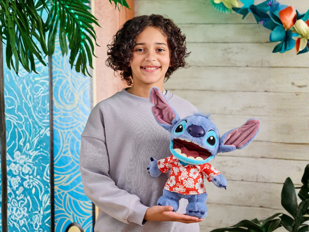 Zabawka interaktywna WOW Disney Aloha Stitch DIS-1027-01 Radosne dziecko w szarej bluzie trzyma w ramionach maskotkę Disney Stitcha w koszuli hawajskiej. Tło to stylizowane deski i tropikalne dekoracje.