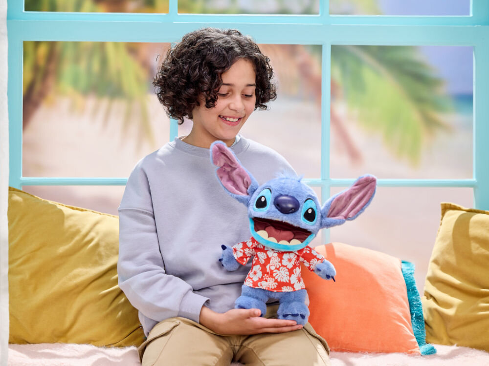 Zabawka interaktywna WOW Disney Aloha Stitch DIS-1027-01 Uśmiechnięte dziecko w jasnych spodniach i szarej bluzie siedzi na kanapie przy oknie z tropikalnym widokiem, trzymając maskotkę Disney Stitcha.