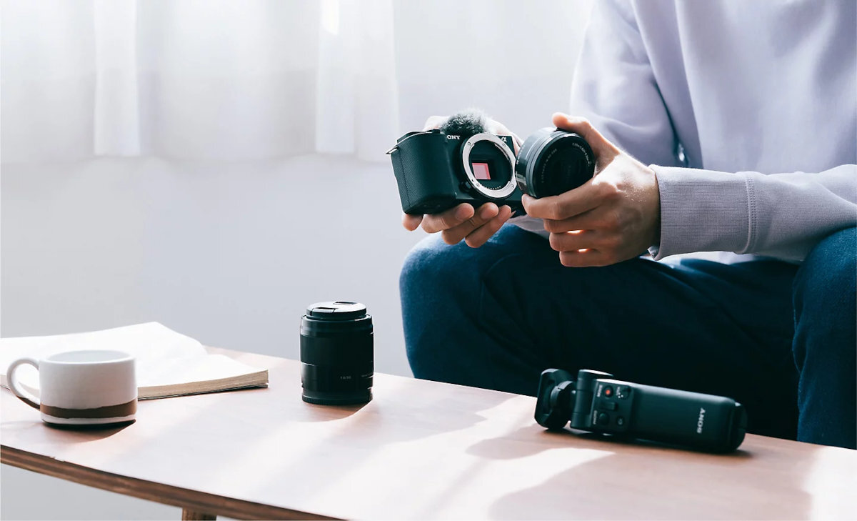 SONY ZV-E10LBD filmy 4K 6K zdjęcia o rozdzielczości 24,2 MP ZV-E10 twórcom hybrydowym wideo fotografią - Osoba montująca wymienny obiektyw do bezlusterkowego aparatu fotograficznego na drewnianym stoliku z akcesoriami i filiżanką kawy