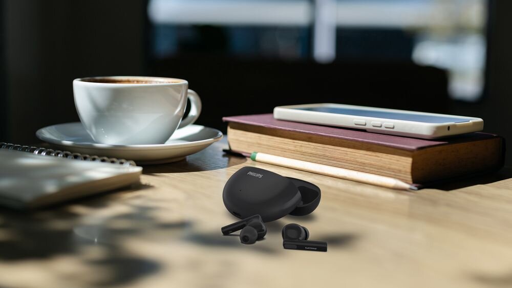 Na drewnianym stole znajdują się czarne słuchawki dokanałowe z etui marki PHILIPS, obok filiżanki kawy, książki, ołówka i smartfona.