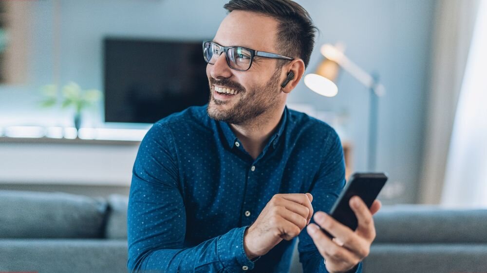 Mężczyzna w okularach siedzi na kanapie, trzymając w ręku smartfon. W uchu ma czarne słuchawki dokanałowe.