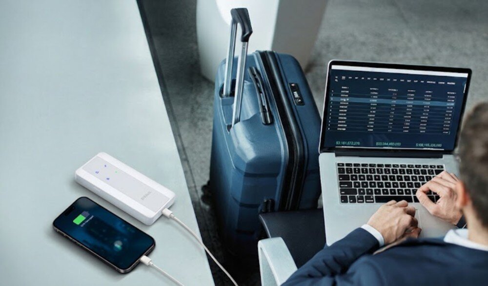 Router D-LINK F518 Mężczyzna siedzący przy laptopie obok walizki, przy którym na blacie leży białe urządzenie połączone kablem z ładującym się smartfonem