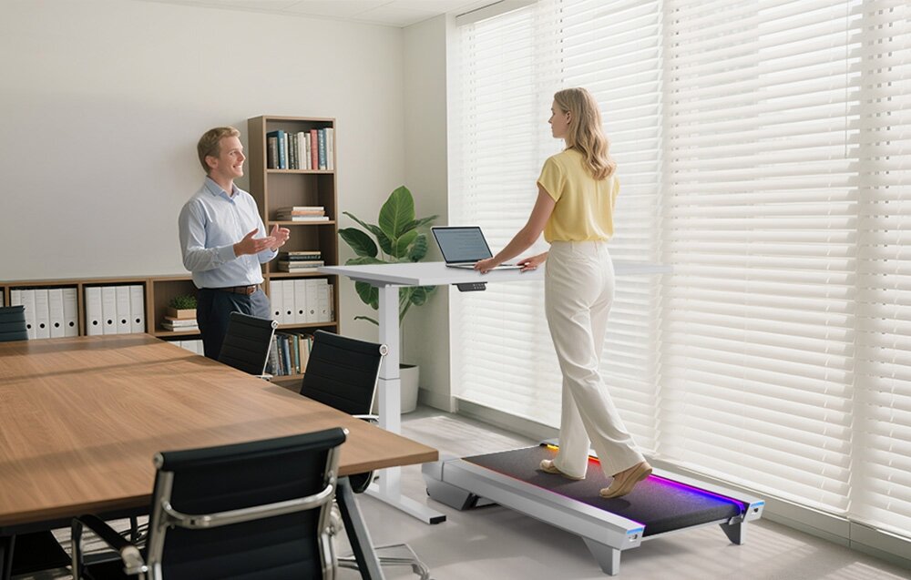 Bieżnia elektryczna UREVO CyberPad for Office 550W 6 km-h pomieszczenie biuro osoba pracuje przy biurku chodząc na bieżni rozmawi z osoba stojącą obok bieżni walking pad aktywności przy biurku spacery smukła konstrukcja (ok. 16,3 cm wysokości) pas biegowy 42 × 100 cm naturalny i komfortowy ruch