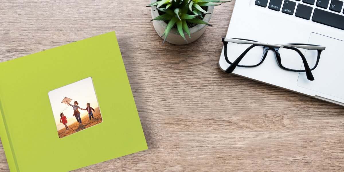 Album HAMA Fine Art Jumbo Białe kartki Zielony (100 stron) Zielony album fotograficzny z okienkiem na własne zdjęcie, leżący na drewnianym biurku obok laptopa, okularów i rośliny doniczkowej.