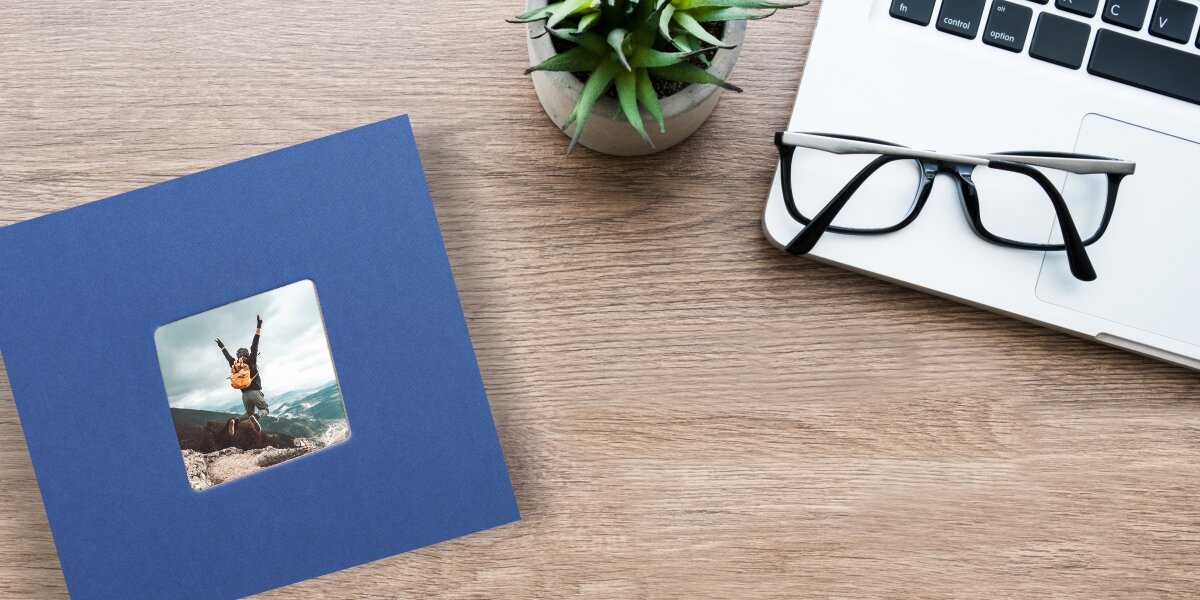 Widok z góry na niebieski album fotograficzny leżący na drewnianym biurku obok laptopa, okularów i rośliny doniczkowej.