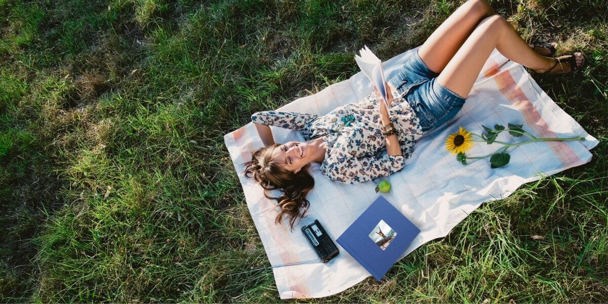 Młoda kobieta czytająca książkę na kocu w ogrodzie, obok której leży niebieski album fotograficzny Hama.