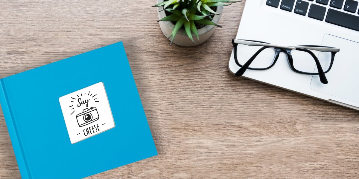 Album HAMA Fine Art Jumbo Białe kartki Błękitny (100 stron) Błękitny album fotograficzny z okienkiem na własne zdjęcie, leżący na drewnianym biurku obok laptopa, okularów i rośliny doniczkowej.