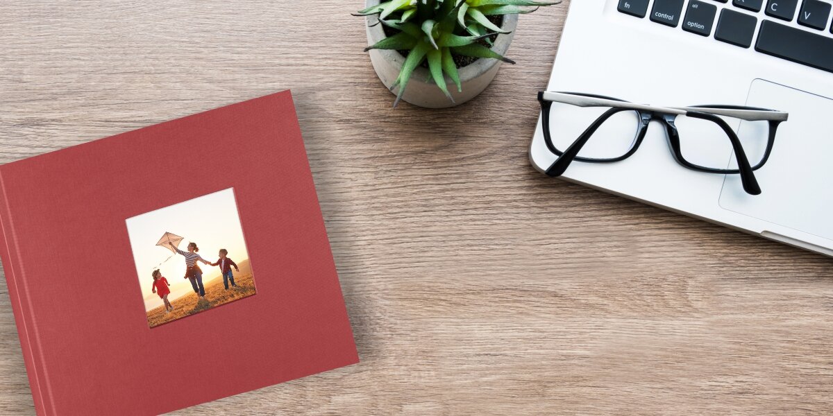 Album HAMA Fine Art Jumbo Białe kartki Bordowy (100 stron) Bordowy album fotograficzny z okienkiem na własne zdjęcie, leżący na drewnianym biurku obok laptopa, okularów i rośliny doniczkowej.