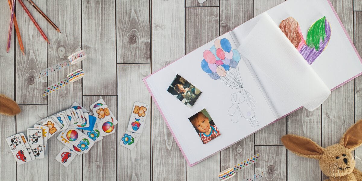 Album HAMA Joana Różowy (50 stron) Wnętrze dziecięcego albumu z białymi kartami, wklejonymi zdjęciami dzieci i kolorowymi rysunkami, otoczone przyborami plastycznymi i zabawkami.