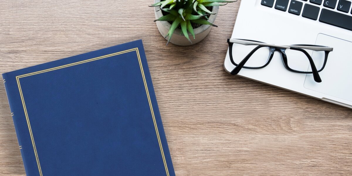 Album HAMA London Super Jumbo Białe Kartki Niebieski (80 stron) Elegancki niebieski album na zdjęcia ze złotymi zdobieniami leżący na drewnianym biurku obok laptopa, okularów i rośliny doniczkowej.