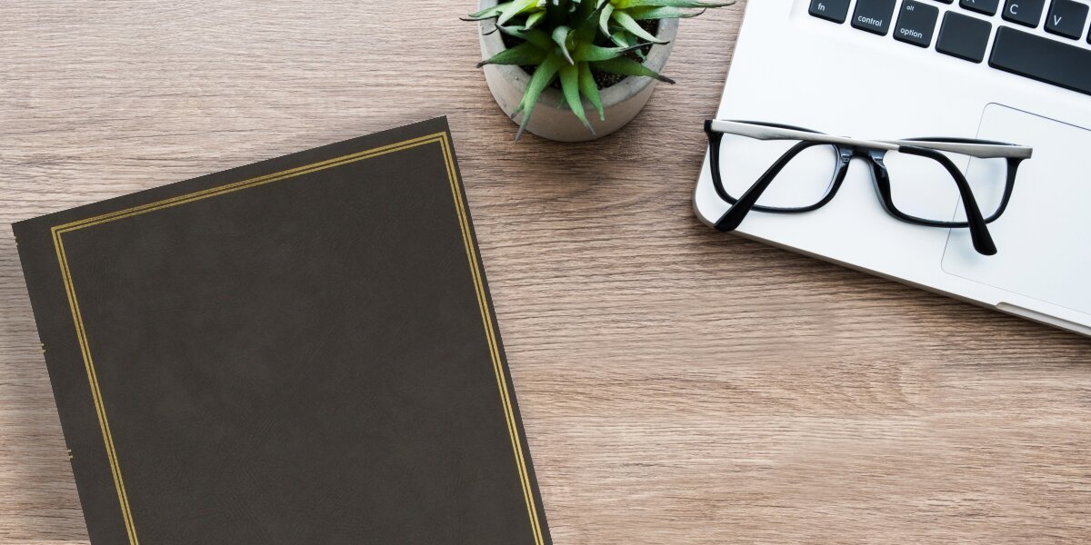 Na drewnianym blacie znajduje się szary album z delikatnym złotym obramowaniem, obok leży para okularów, laptop i mała roślina w doniczce.