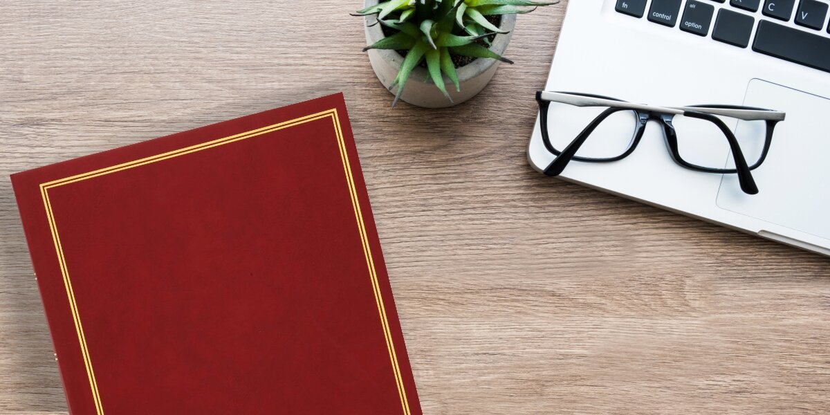 Album HAMA London Super Jumbo Białe Kartki Bordowy (80 stron) Elegancki Bordowy album na zdjęcia ze złotymi zdobieniami leżący na drewnianym biurku obok laptopa, okularów i rośliny doniczkowej.