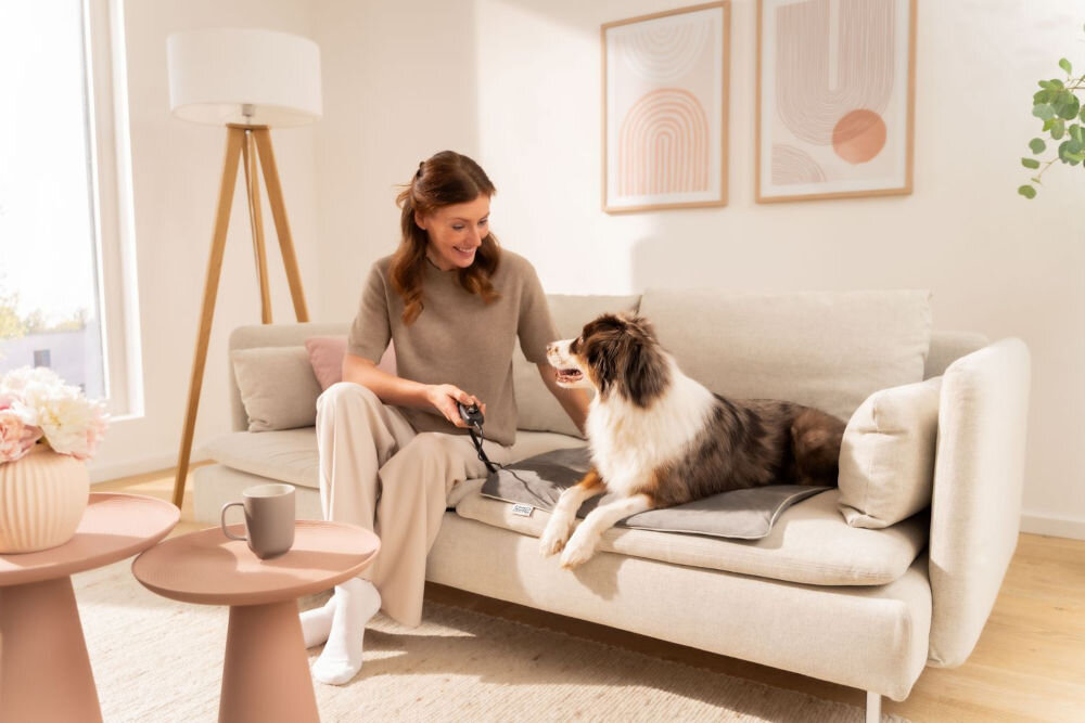 Mata podgrzewana BEURER Sammy Kobieta siedzi na beżowej kanapie obok psa rasy Australian Shepherd (lub podobnej) leżącego na szarej podgrzewanej macie dla zwierząt. Kobieta trzyma w ręce kontroler do maty. W tle nowoczesna lampa i obrazy na ścianie.