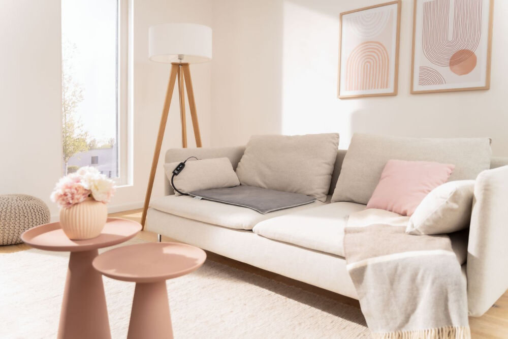 Mata podgrzewana BEURER Sammy Zdjęcie prezentujące pustą szarą podgrzewaną matę dla zwierząt umieszczoną na beżowej kanapie. Obok maty leży mała poduszka. Widoczny jest też kontroler maty. W tle nowoczesny wystrój: lampa i stoliki w kolorze różowym.