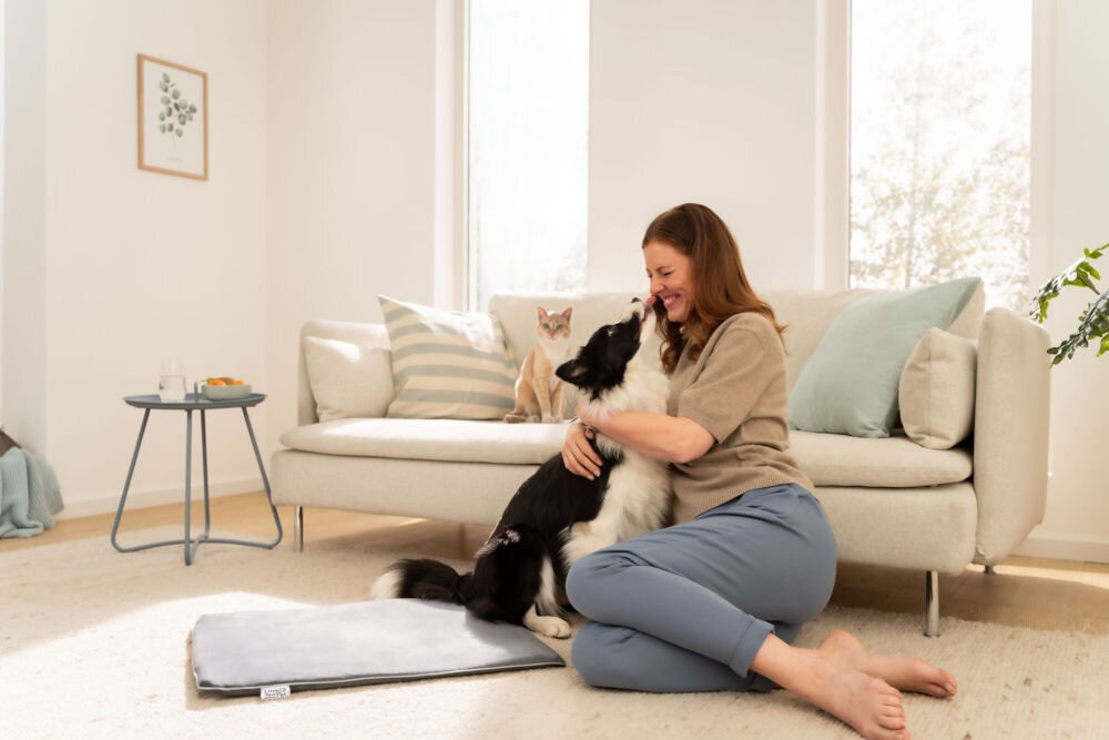 Mata podgrzewana BEURER Sammy Kobieta siedzi na jasnym dywanie i obejmuje czarno-białego psa (Border Collie). Pies podskakuje, aby ją polizać. Obok leży szara podgrzewana mata, a na kanapie w tle siedzi kot.