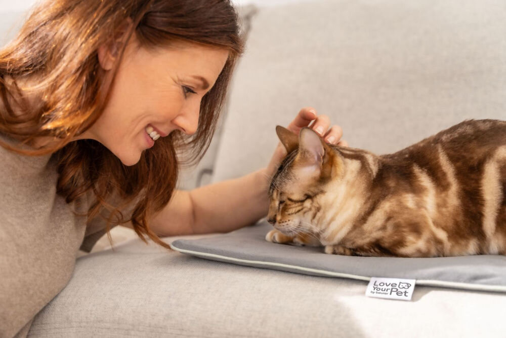 Mata podgrzewana BEURER Lilli Zbliżenie na uśmiechniętą kobietę delikatnie gładzącą brązowo-cynamonowego kota (Bengal) po głowie. Kot leży na szarej podgrzewanej macie, na której widoczna jest metka z napisem Love Your Pet
