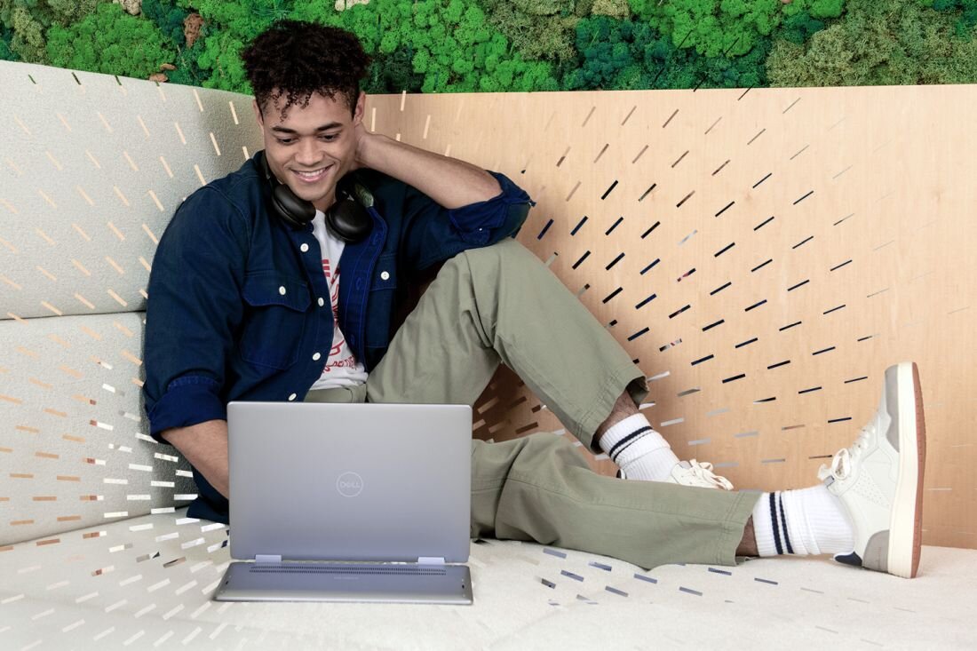 Osoba siedzi na kanapie z laptopem marki Dell na kolanach. W tle widoczna jest ściana z zielonymi roślinami.