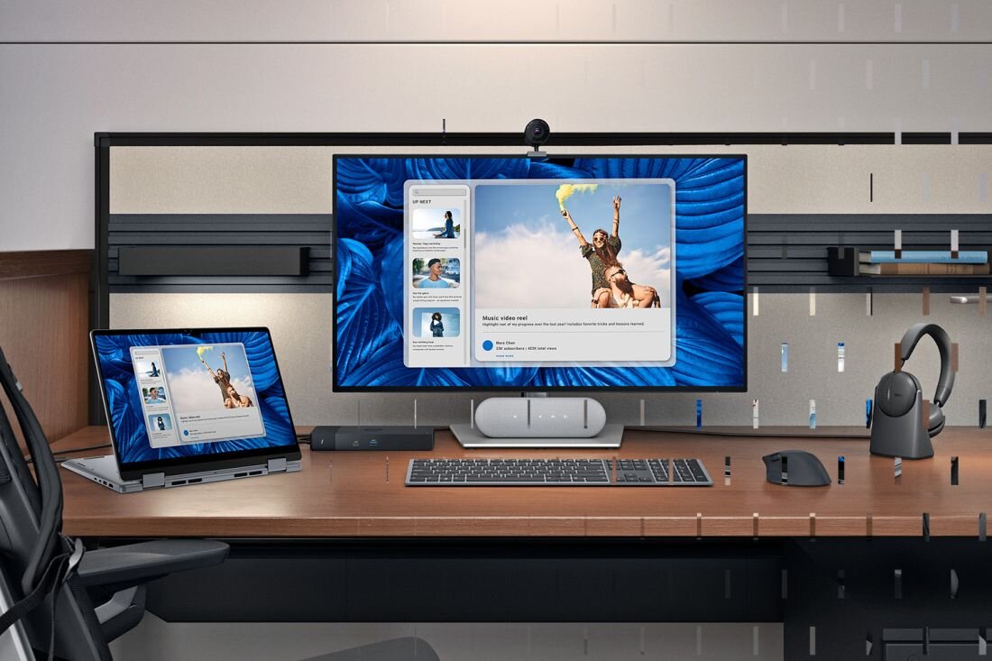 Na biurku znajduje się laptop oraz monitor, oba wyświetlają ten sam obraz przedstawiający osobę z uniesionymi rękami na tle nieba. Obok monitora znajdują się klawiatura, myszka i słuchawki.