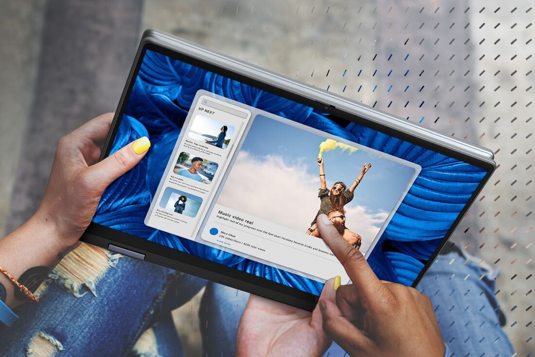 Osoba trzyma laptop w trybie tabletu, na ekranie widoczny jest interfejs z wideo i listą odtwarzania. W tle widać niebieski materiał, a osoba ma pomalowane na żółto paznokcie.