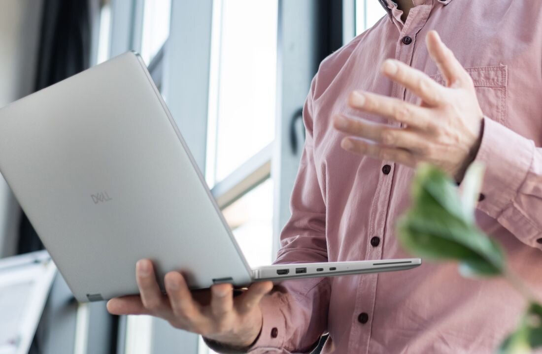 Osoba trzyma otwarty laptop z widocznym logo 'Dell', ubrana w różową koszulę. W tle widać okno i roślinę.