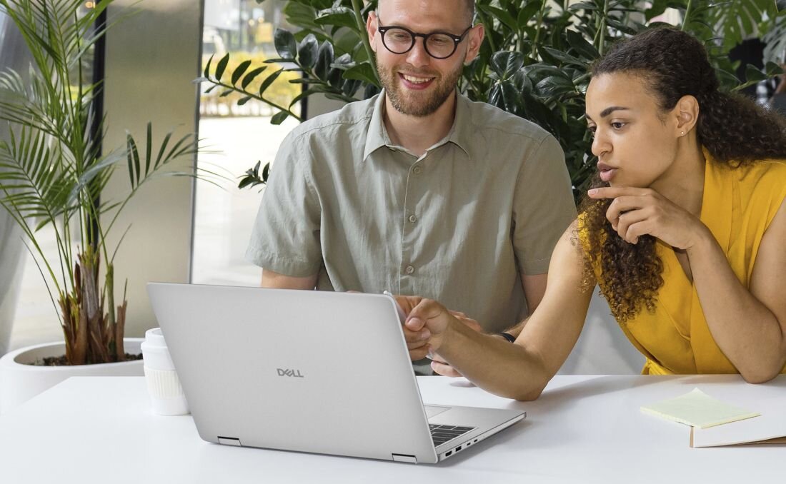 Dwie osoby siedzą przy stole, patrząc na otwarty laptop z logo 'Dell'. W tle widoczne są rośliny doniczkowe.